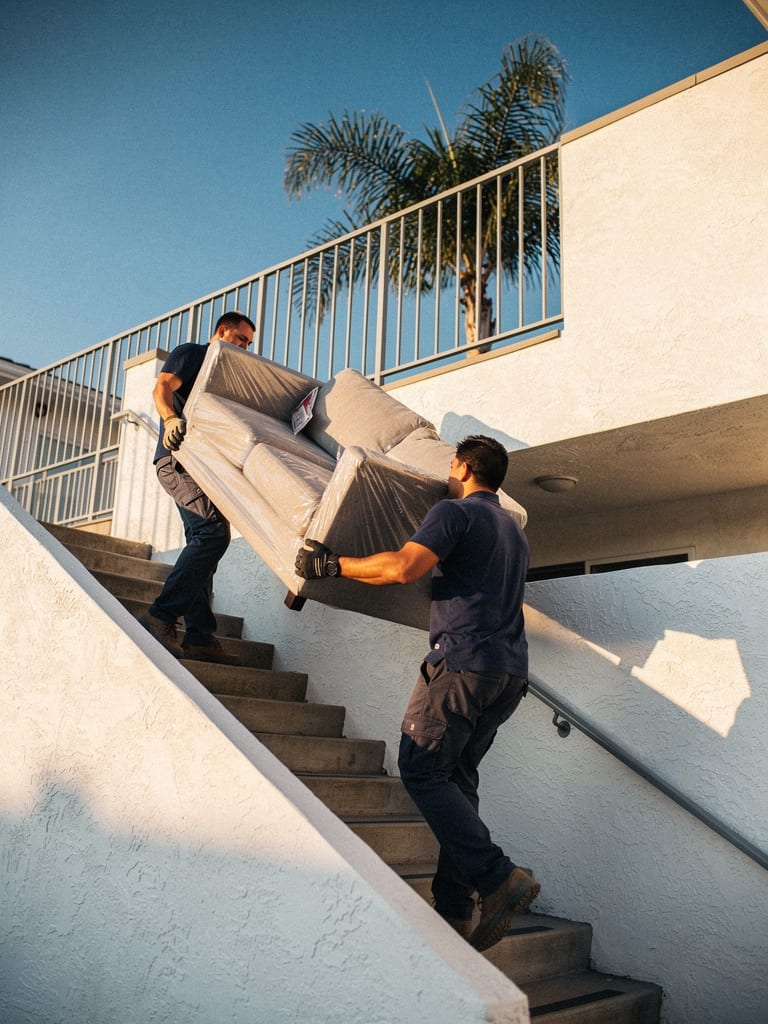 Two delivery workers carry a wrapped sofa up the outdoor stairs of an apartment building